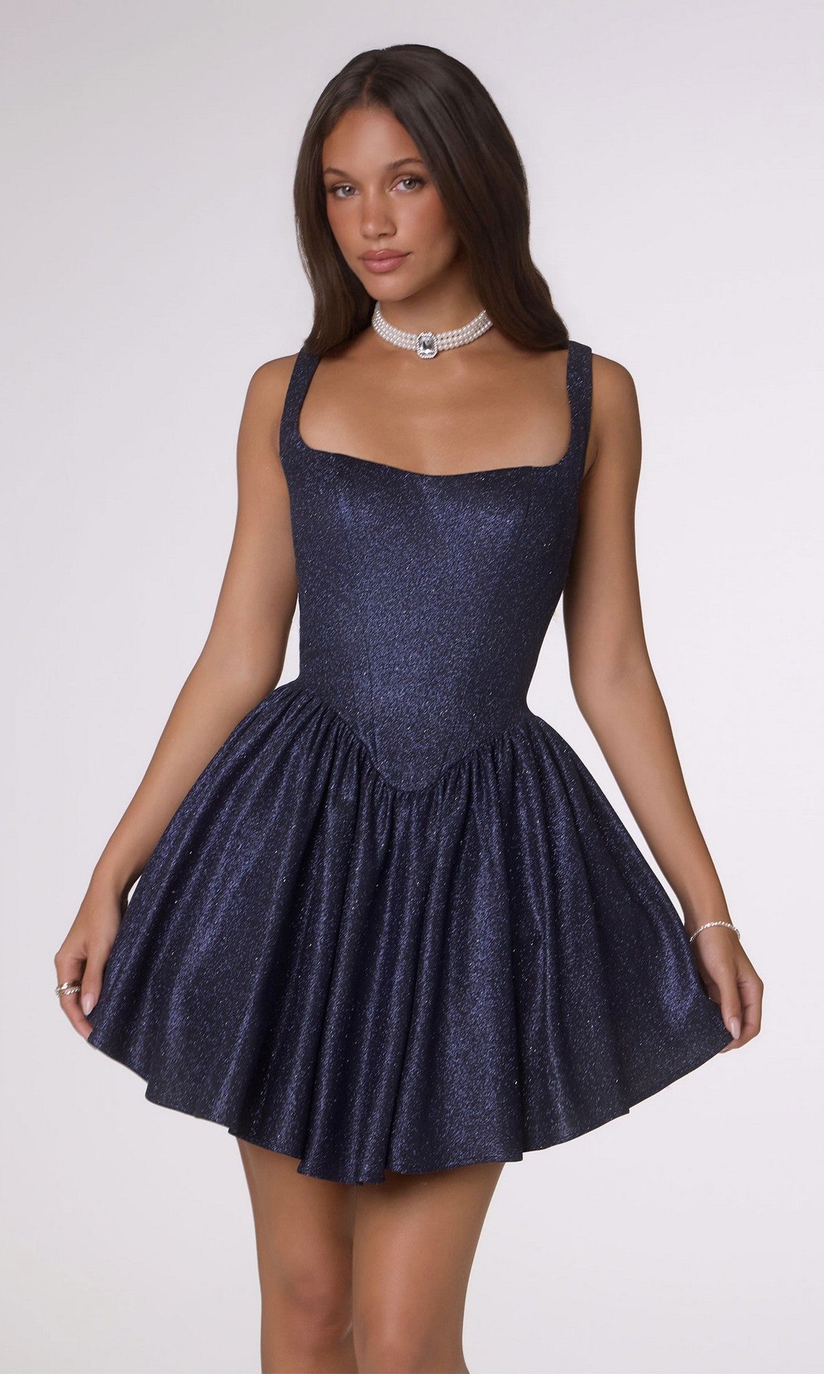 A woman models the Amoris T738 short navy homecoming dress with a fit-and-flare silhouette and shimmering stretch fabric, paired with a pearl choker necklace, against a plain light background.